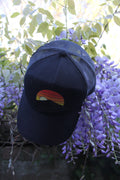 Casquette trucker rétro noire « Terril au coucher de soleil » avec visuel terril stylisé, posée dans des fleurs violettes en extérieur