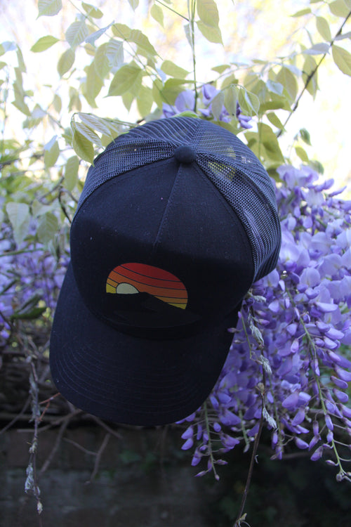Casquette trucker rétro noire « Terril au coucher de soleil » avec visuel terril stylisé, posée dans des fleurs violettes en extérieur